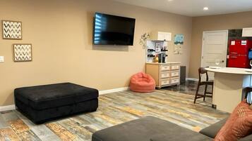 A living room with a flat-screen TV, a sofa, a bean bag, and a kitchen area with a refrigerator and cabinets.