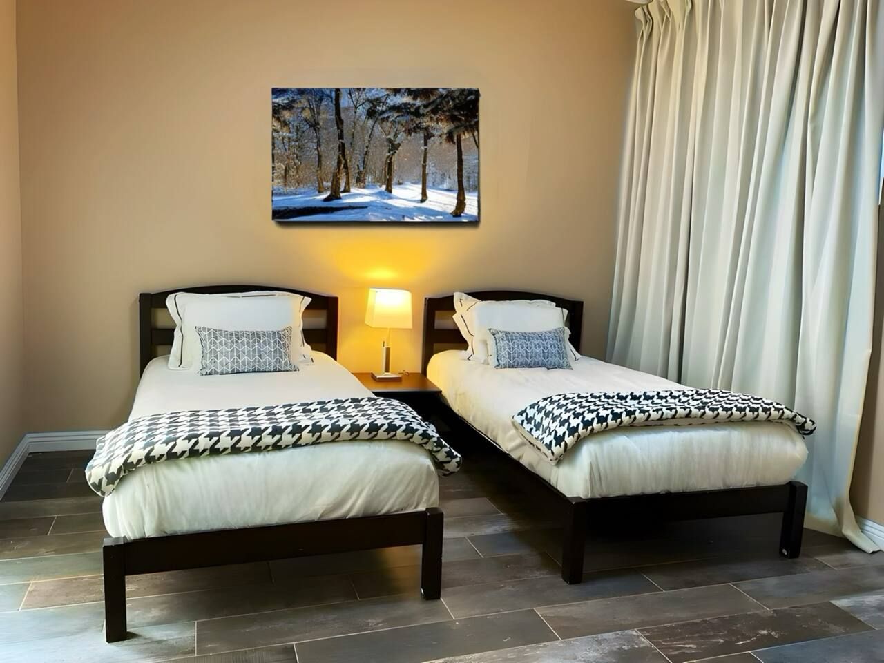 A hotel room with two beds, a nightstand with a lamp, a painting of a snowy forest, and a large window with curtains.
