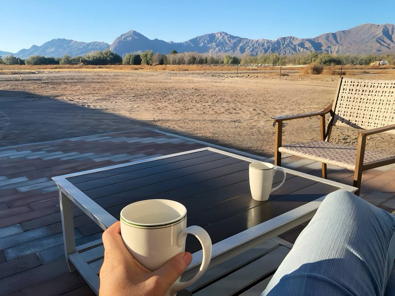 Une personne assise sur une chaise longue, devant une petite table, tenant une tasse à la main, et une deuxième tasse posée sur la table.