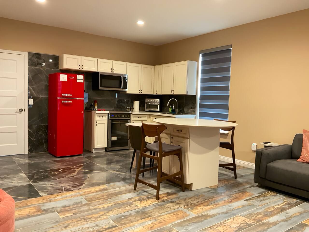 A compact kitchen with a red refrigerator, a microwave, and a built-in oven. There is a small island with a chair, and a sofa is visible in the living area.