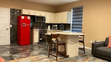 A compact kitchen with a red refrigerator, a microwave, and a built-in oven. There is a small island with a chair, and a sofa is visible in the living area.