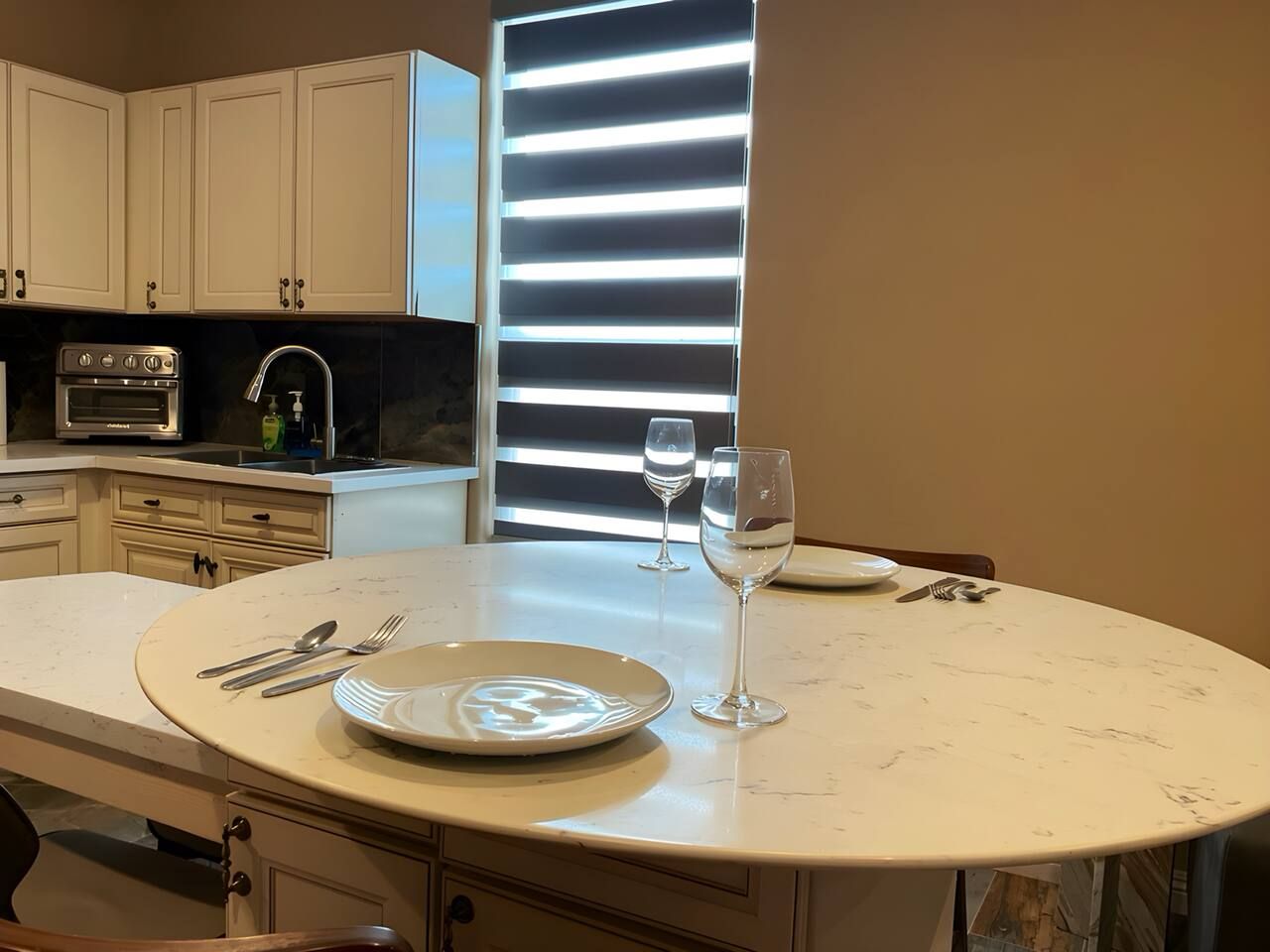 Une cuisine avec une table à manger ronde, un four encastré et une fenêtre avec des stores.