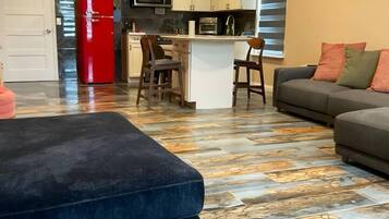 Cozinha moderna com armários brancos, uma geladeira vermelha e um micro-ondas. Há uma área de jantar com um banco de bar e um sofá com almofadas. O chão tem um design de piso em mosaico.