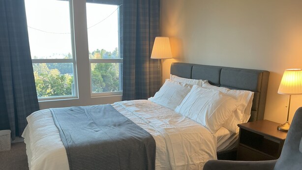 Cama bem arrumada com roupa de cama branca e manta cinza, uma poltrona cinza e uma mesa de cabeceira com abajur.