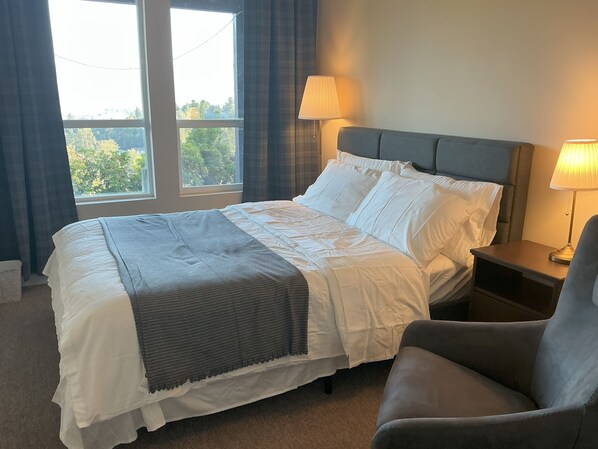 A neatly made bed with white linens and a gray throw, a gray armchair, and a nightstand with a lamp. - Bright Entire Walkout Suite, Stunning Mt Baker Views (Abbotsford)