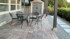 Um alpendre coberto com piso de tijolos, uma mesa com cadeiras e um galpão ao fundo.
