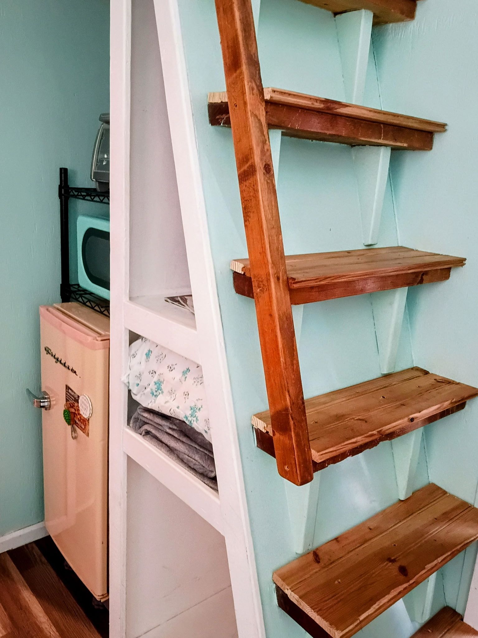 Habitación con un armario blanco, un microondas encima y una estantería de madera apoyada en una pared azul claro.