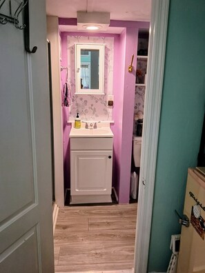 Une petite salle de bain avec un lavabo blanc, un miroir et une porte donnant sur une autre pièce.