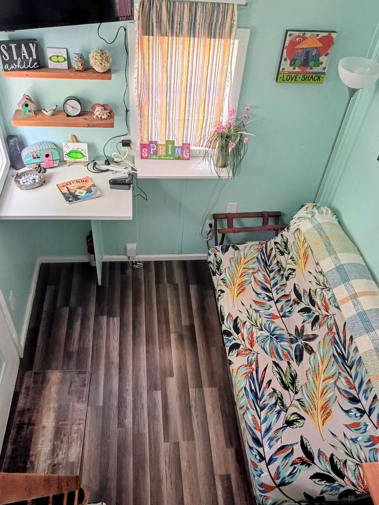 Una habitación pequeña con piso de madera, una cama con colcha estampada de flores, un escritorio pequeño con varios objetos, una ventana con cortina a rayas y una pared con un letrero que dice 'LOVE-SHACK'.
