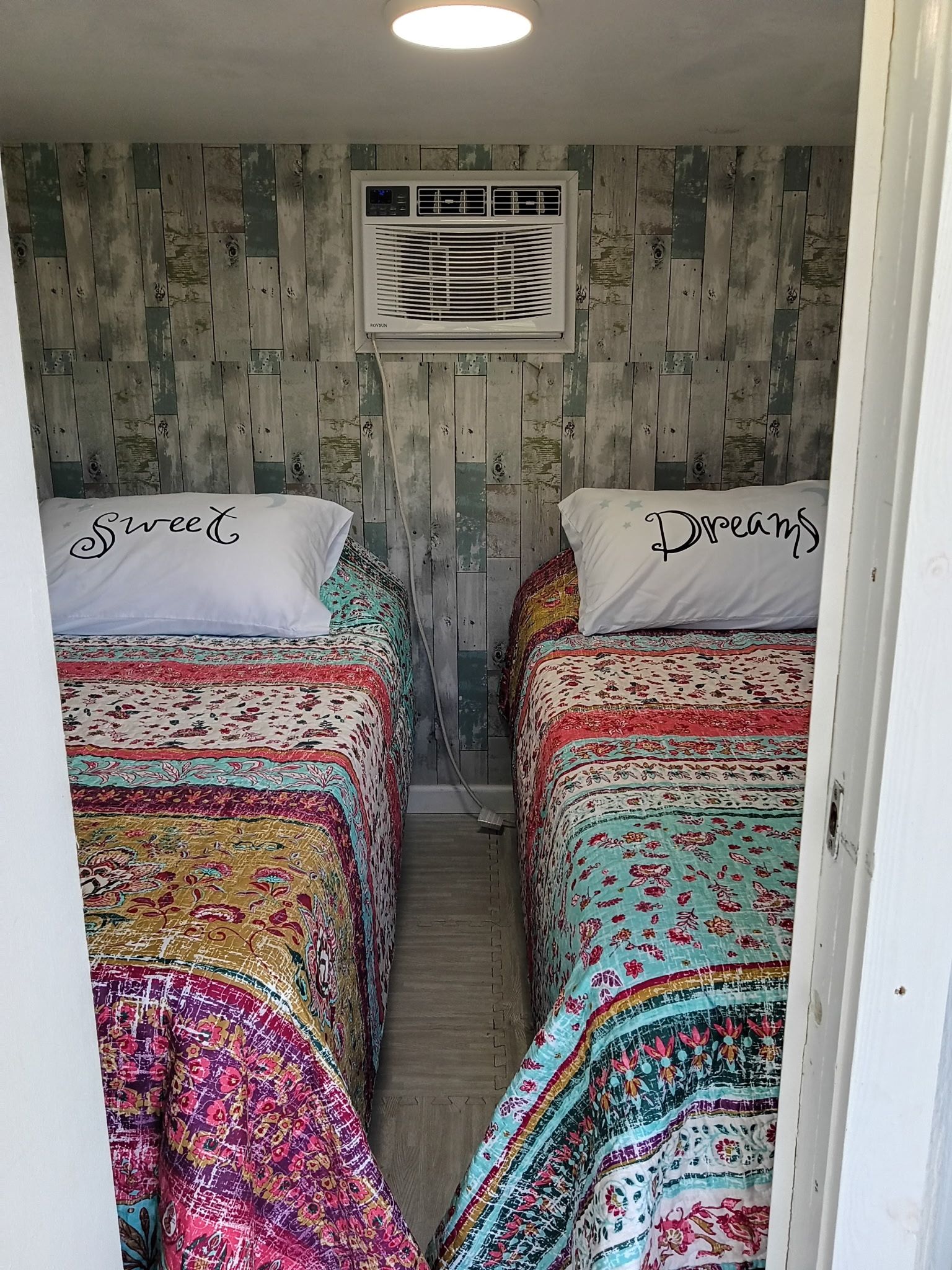 A narrow room with two beds, each with a pillow labeled 'Sweet' and 'Dreams.' An air conditioning unit is mounted on the wall above the beds.