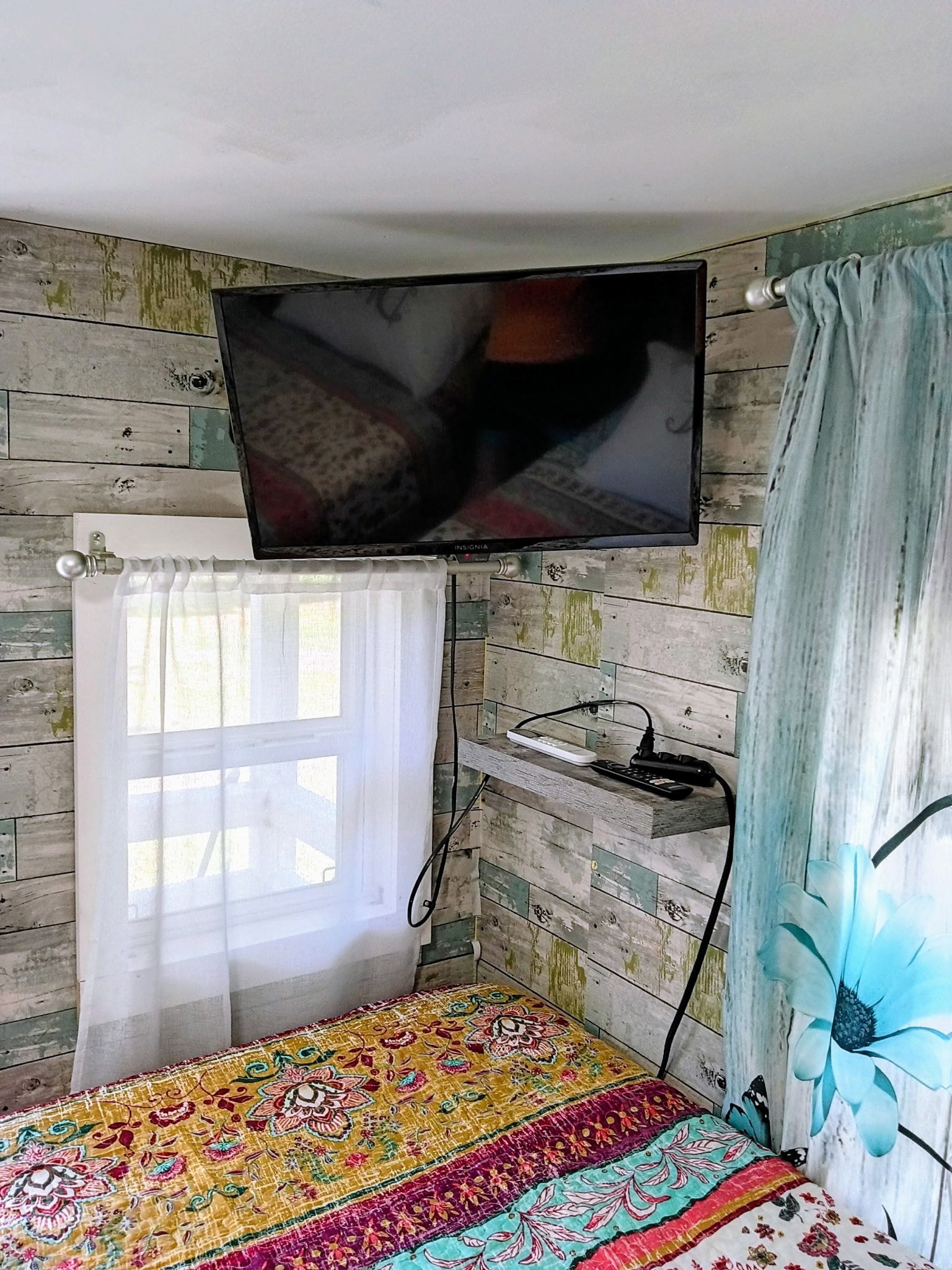 A room with a bed, a television mounted on the wall, a window with a curtain, and a wall with a wooden panel design.