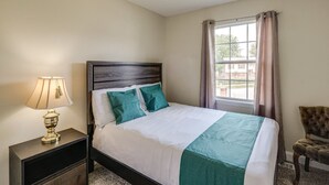 Un lit bien fait, recouvert d’une couverture blanche et turquoise, avec une tête de lit en bois foncé, une lampe de chevet et une chaise placée à droite.