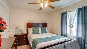 Une chambre avec une tête de lit en bois, un lit avec une literie blanche et turquoise, une table de chevet avec une lampe, une commode et un ventilateur de plafond.