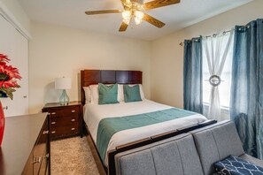 Un dormitorio con cabecera de madera, cama con ropa de cama blanca y azul turquesa, mesita de noche con lámpara, cómoda y ventilador de techo.