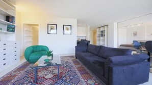 Ein modernes Wohnzimmer mit einem blauen Sofa, einem grünen Sessel, einem Glastisch und einem gemusterten Teppich. Im Hintergrund sind Bücherregale und ein Essbereich zu sehen.