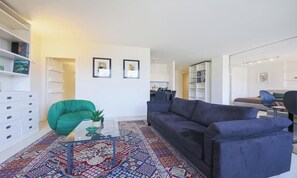 A modern living room with a blue sofa, a green chair, a glass coffee table, and a patterned rug. There are bookshelves and a dining area in the background. - VASARI LUXURY – SOPHISTICATED FLORENCE HOME WITH RIVER VIEWS (Firenze)