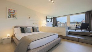Ein modernes Schlafzimmer mit einem großen Bett, Nachttischen, einer Kommode und Blick auf die Stadt durch große Fenster.