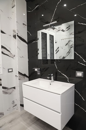 A modern bathroom with a large white sink, a mirror, and black marble walls. - Vrbo Property (Lido Manfria)