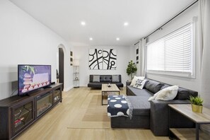 Ein modernes Wohnzimmer mit Flachbildfernseher, einer Couch, einem Couchtisch und einem großen Fenster mit Jalousien.