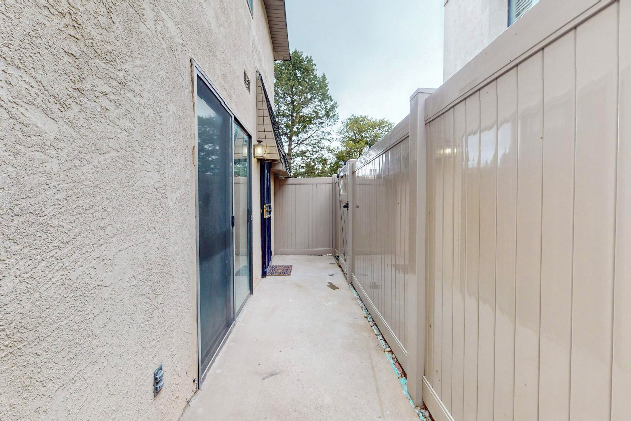 A narrow concrete pathway flanked by a beige wooden fence and a textured wall, leading to a door with a glass panel.