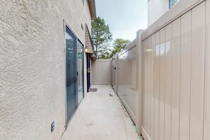 A narrow concrete pathway flanked by a beige wooden fence and a textured wall, leading to a door with a glass panel.
