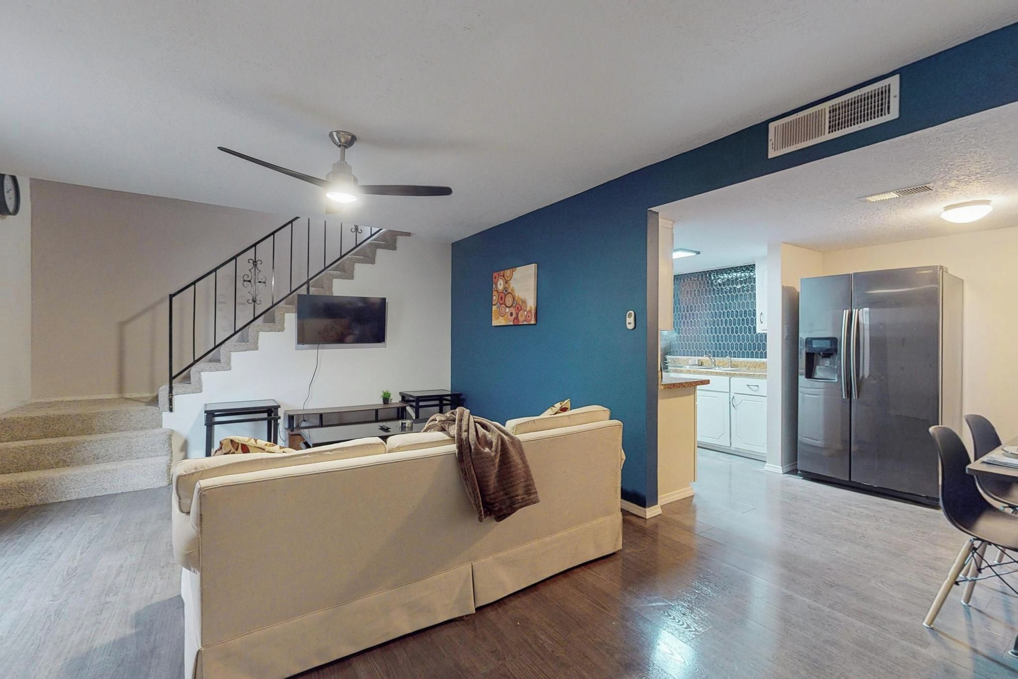 A modern living room with a sofa, a dining area, a kitchen with a refrigerator, and a staircase.