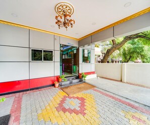 Entrada de hotel con fachada roja y blanca, un letrero de bienvenida, piso con diseño y un árbol afuera.