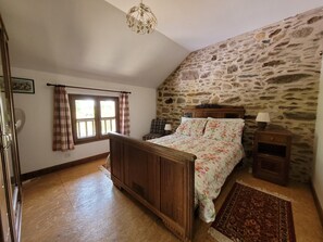 A bedroom with a stone wall, a wooden bed, a nightstand, a lamp, and a window with curtains. - Authentic Estate in the Dordogne With a Pool (Saint-Saud-Lacoussiere)