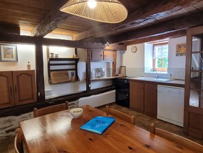 A rustic kitchen with wooden beams, a dining table with a bowl and a blue folder, and a window with a view of greenery. - Authentic Estate in the Dordogne With a Pool (Saint-Saud-Lacoussiere)