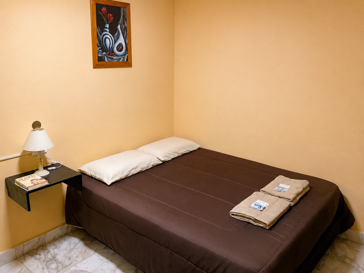 Een eenpersoonsbed met een bruine matras, twee kussens, een nachtkastje met een lamp en een schilderij aan de muur.
