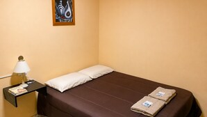 Een eenpersoonsbed met een bruine matras, twee kussens, een nachtkastje met een lamp en een schilderij aan de muur.