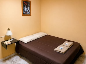 Uma cama de solteiro com colchão castanho, duas almofadas, uma mesa de cabeceira com abajur e um quadro na parede.