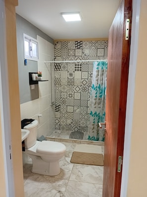 Casa de banho com sanita, chuveiro com azulejos estampados e porta com puxador de metal.