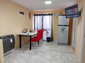 A room with a dining table, red chair, potted plant, refrigerator, and a mounted TV displaying a streaming service. - GENESIS APARTMENT (Puerto San Julian)