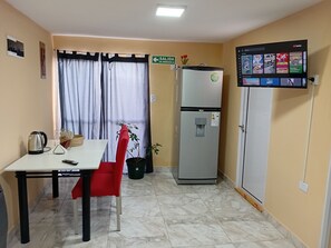 A compact kitchenette with a refrigerator, a water dispenser, a small table with a chair, and a television mounted on the wall displaying various channels.