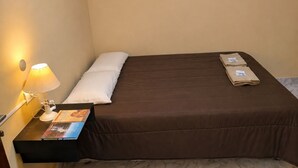 Een enkel bed met een bruine matras en wit kussen, een nachtkastje met een lamp, en een klein, ingelijst fotootje aan de muur.