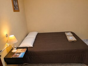 Uma cama de solteiro com colchão marrom e travesseiro branco, uma mesa de cabeceira com abajur, e uma pequena moldura com uma imagem na parede.