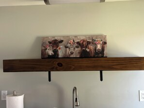 Una cocina con un estante de madera que sostiene un cuadro de vacas, un dispensador de toallas de papel y un grifo.