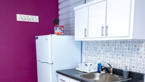 Eine kleine Küche mit weißen Schränken, einer Edelstahl-Spüle und einer violetten Wand.