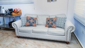 Ein weißes Sofa mit gemusterten Kissen, ein kleiner Tisch mit Küchenutensilien und ein an der Wand befestigtes Schild.