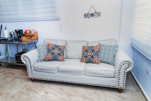 A white sofa with patterned cushions, a small table with kitchen items, and a wall-mounted sign. - Cozy 1BR Getaway in Beautiful Aguada,PR AC & Comfort (Aguada)