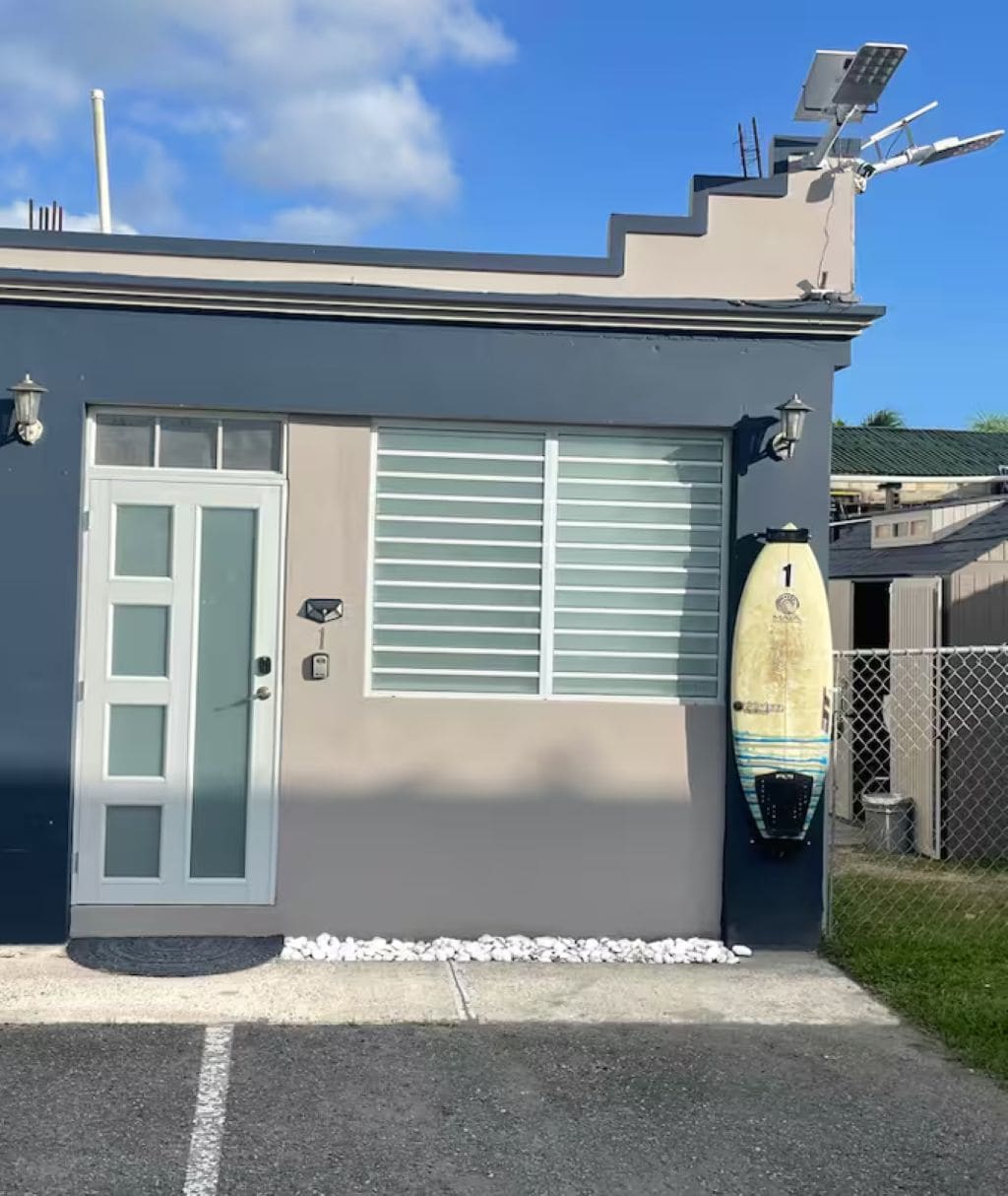 Un edificio con una puerta de garaje, una tabla de surf apoyada en la pared y una valla al fondo.