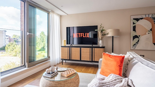 Un salon moderne doté d’une grande fenêtre, d’un téléviseur à écran plat et d’un canapé agrémenté d’un coussin orange.