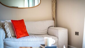 Un salon avec un canapé, un coussin rouge, une tasse blanche et une pile de livres sur une table d’appoint.