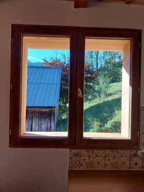 A wooden-framed window with a view of a blue-roofed building and a green landscape. - Menziesii Vacation Rental. In a traditional farmhouse just 5 minutes from the village center (Abondance)