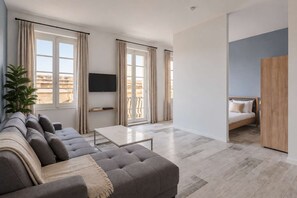 Uma sala de estar moderna com um sofá cinzento, uma mesa de centro e uma TV em um móvel. Há uma varanda com cortinas e uma planta.