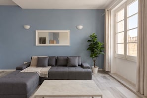 A living room with a grey sofa, a white coffee table, a large window with curtains, and a potted plant. - Cozy Flat with Rooftop in the Heart of Valletta (Valletta)