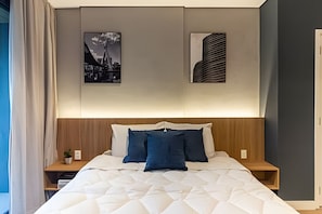 A neatly made bed with a quilted cover, two pillows, and a wooden headboard. There are two bedside tables with lamps, and the walls are adorned with two framed pictures. - Nato Itaim (São Paulo)