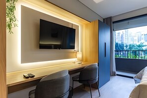 Een moderne hotelkamer met een flatscreen televisie aan een muur, een houten bureau met twee stoelen, en een groot raam met uitzicht op de stad.
