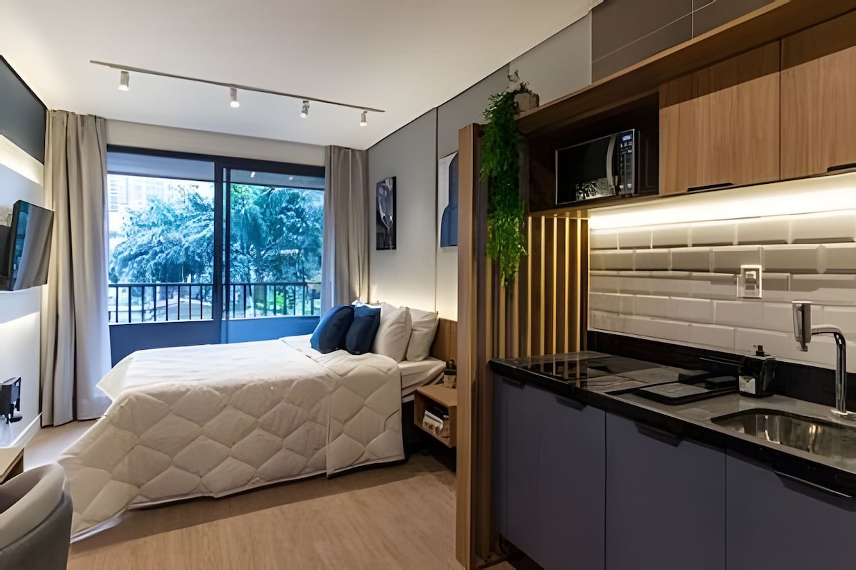 A modern hotel room with a bed, a kitchenette, and a large window offering a view of trees.